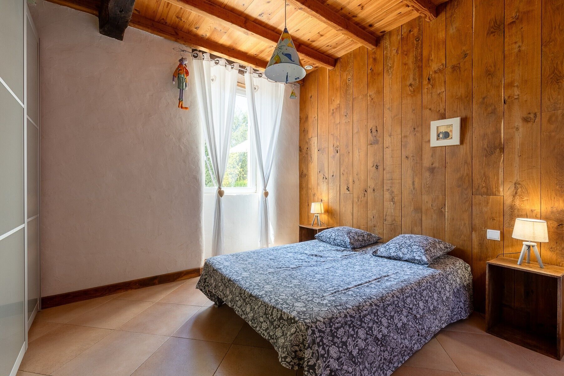 A bedroom with a wooden ceiling, a bed with patterned bedding, a nightstand with a lamp, and a window with curtains.