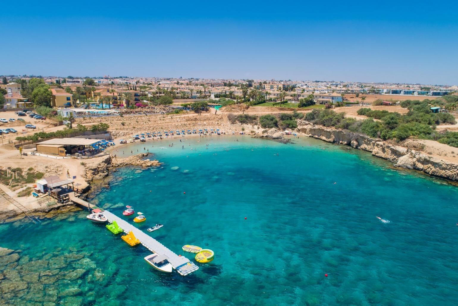 Vista aérea de una zona costera con aguas turquesas cristalinas, un muelle con barcos y una playa con sombrillas.