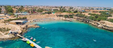 Vista aérea de una zona costera con aguas turquesas cristalinas, un muelle con barcos y una playa con sombrillas.