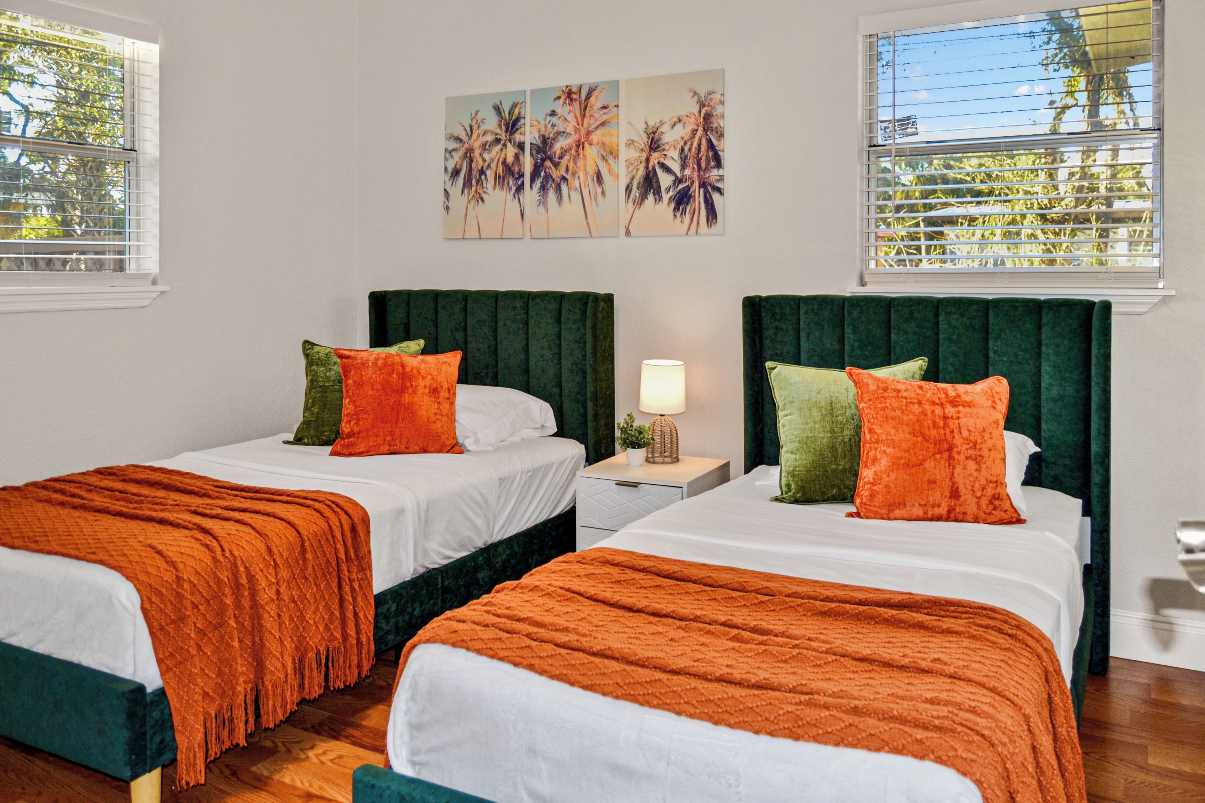 A hotel room with two beds, each with a green headboard, white bedding, and orange throw blankets. There is a nightstand with a lamp and a small plant.