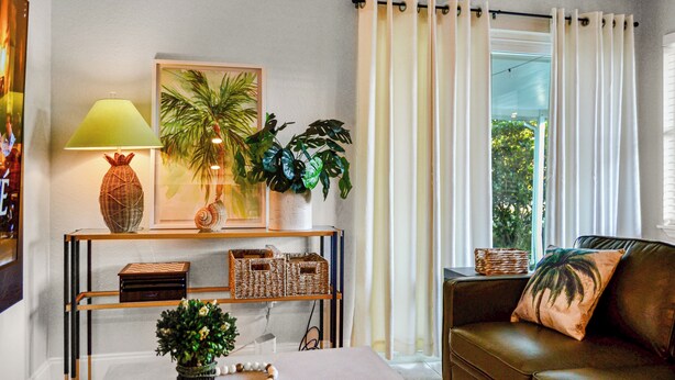 A living room with a sofa, a coffee table, a lamp, a framed picture, a plant, and a window with curtains. - Vibrant Cuban Style 3BR with AC near Beach & Downtown (Fort Myers)