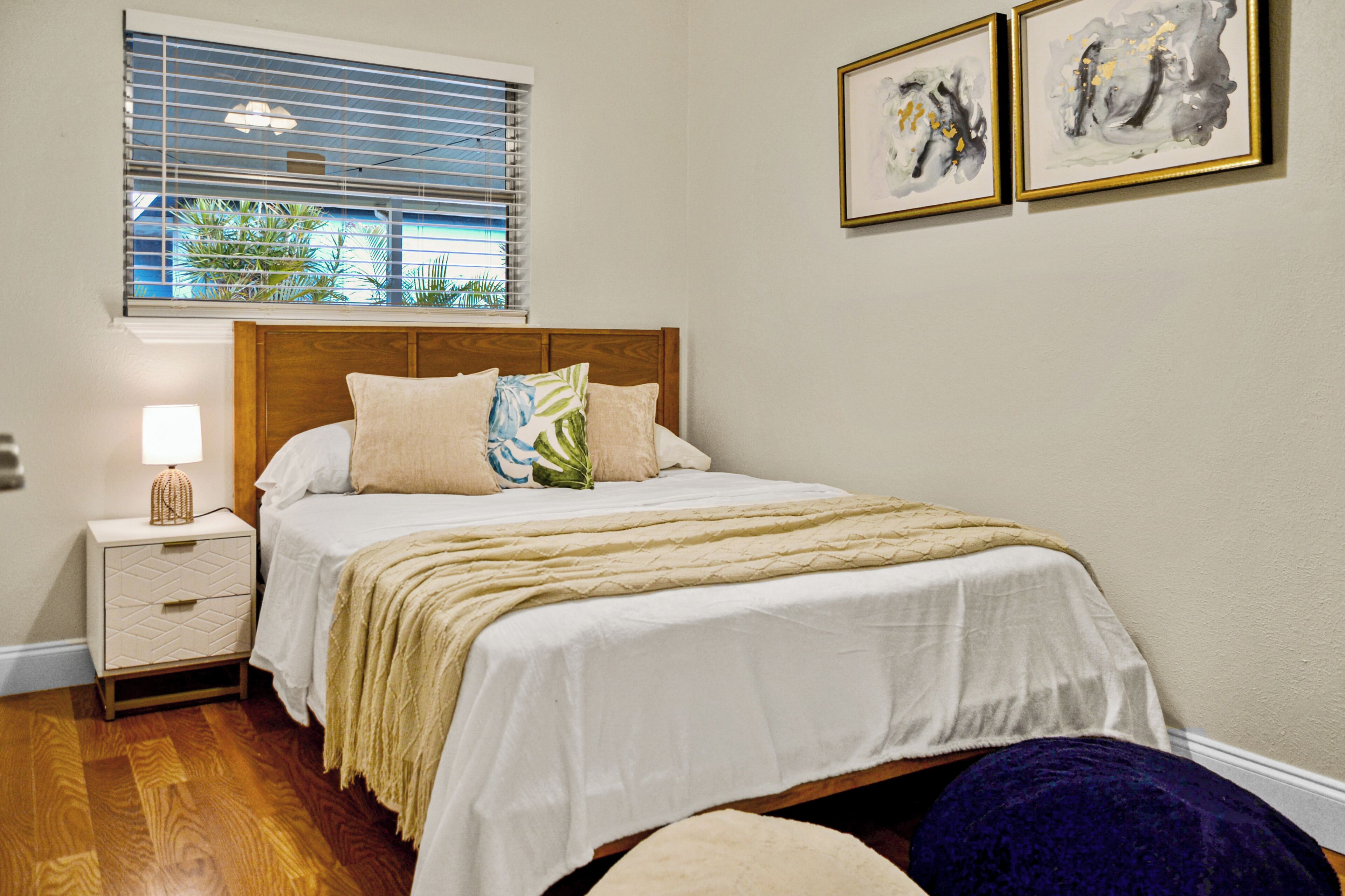 A bedroom with a bed, bedside table, lamp, window with blinds, and two framed artworks on the wall.