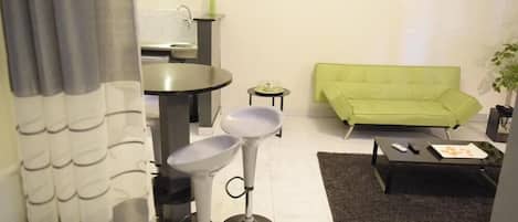 Una sala de estar moderna con un sofá verde, una mesa de comedor redonda, dos taburetes de bar, una mesita auxiliar pequeña y una mesa de centro con controles remotos.