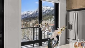 A kitchen with a stainless steel refrigerator, a dining area with a table and chairs, and a balcony with a mountain view. - Elevation Hydeaway at Ascent (Canmore)