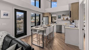 A modern kitchen with a central island, a dining area with bar stools, and a view of mountains through large windows. - Elevation Hydeaway at Ascent (Canmore)