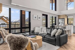 Een moderne woonkamer met een zwarte leren bank, een ronde salontafel en grote ramen met uitzicht op bergen.