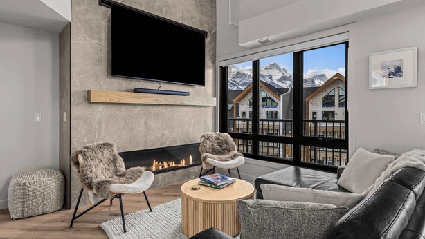 A modern living room with a fireplace, a flat-screen TV, and a large window with a mountain view. - Elevation Hydeaway at Ascent (Canmore)
