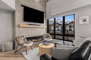 A modern living room with a fireplace, a flat-screen TV, and a large window with a mountain view. - Elevation Hydeaway at Ascent (Canmore)