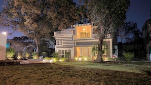 Uma casa moderna de dois andares com um jardim da frente bem iluminado, árvores grandes e um arco visível.
