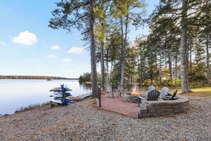 En træterrasse ved søen med Adirondack-stole, bålplads og kanoer ved vandet.