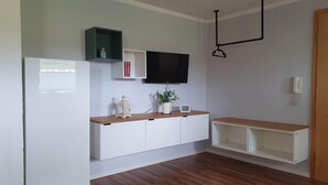 A modern living room with a wooden floor, white cabinetry, a mounted TV, a hanging light fixture, and a small plant on the counter. - 1 room apartment with terrace, quietly situated on the edge of a field. (Remseck am Neckar)