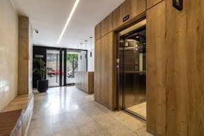 Um hall moderno com uma receção de madeira, um elevador de vidro e um caminho livre para a saída.