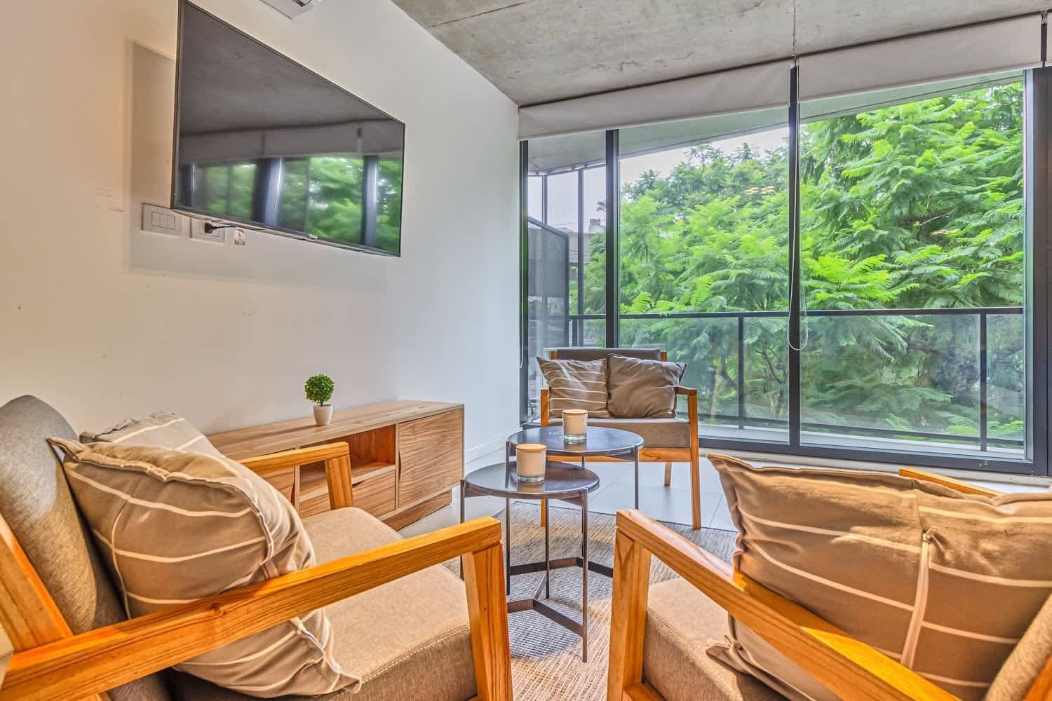 A modern room with a sofa, armchairs, a small table, and a TV. Large windows offer a view of lush greenery outside.