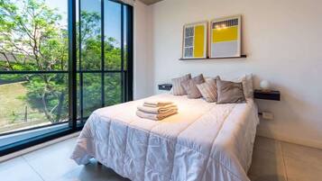 Un dormitorio con un ventanal grande, una cama con cubrecama blanco y dos cuadros abstractos en la pared.