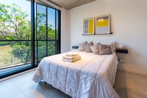 Ein Schlafzimmer mit großem Fenster, einem Bett mit weißer Daunendecke und zwei an der Wand befestigten Kunstwerken.