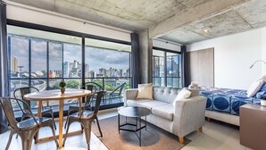 A modern living room with a sofa, a dining table, and chairs, large windows with a city view, and a balcony. - Loft Elegante a 5 Minutos de Puerto Madero 8A (Buenos Aires)