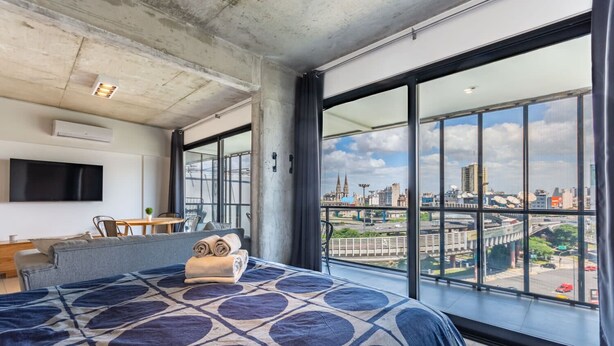 A modern hotel room with a large bed, a TV, a dining area, and a city view through large windows. - Loft Elegante a 5 Minutos de Puerto Madero 8A (Buenos Aires)