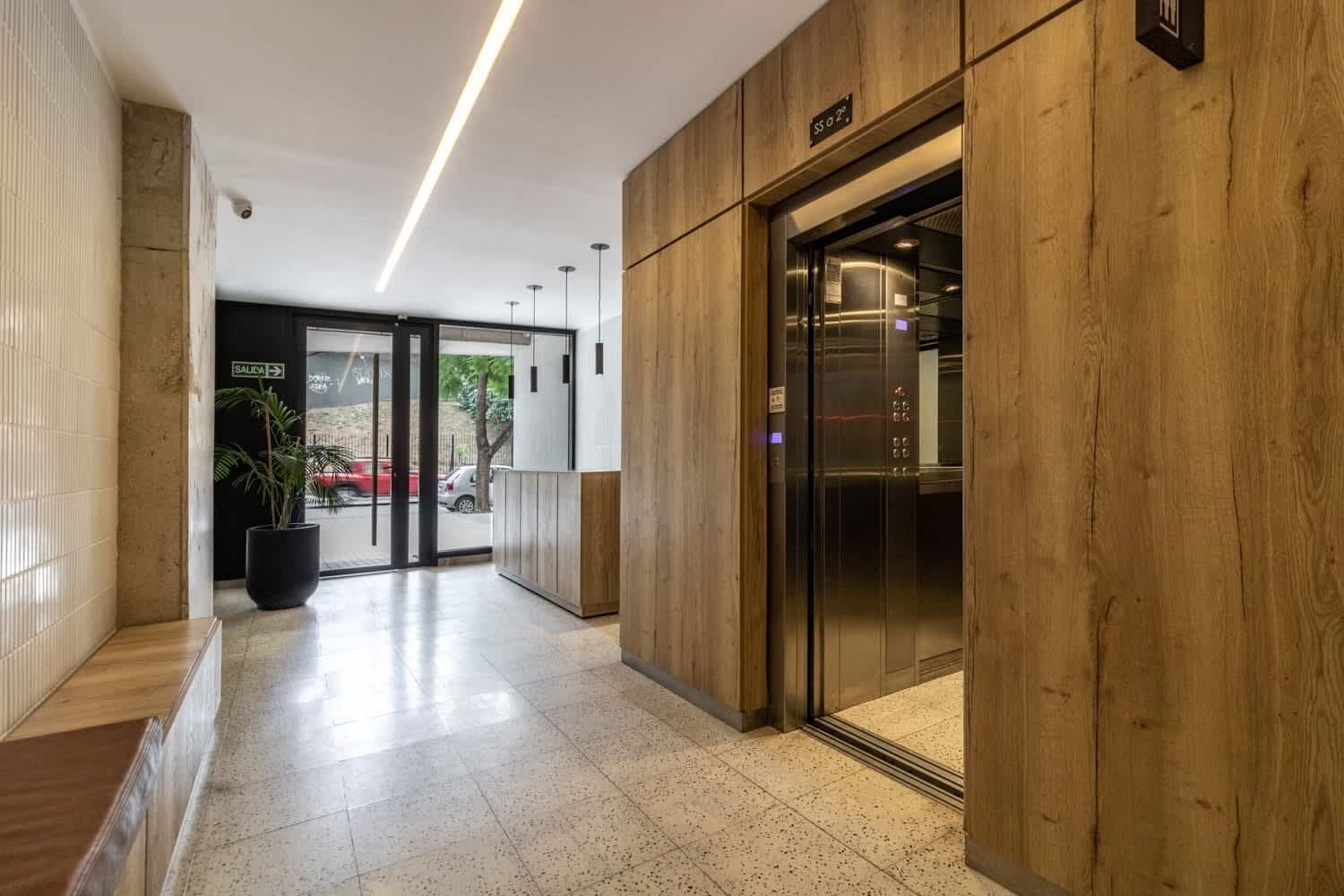 Un vestíbulo moderno con mostrador de recepción de madera, un ascensor de vidrio y un acceso despejado a la salida.
