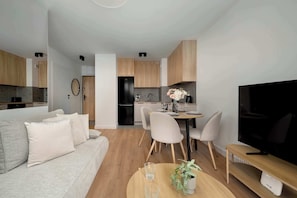 Ein modernes Wohnzimmer mit Sofa, Esstisch, Stühlen, einem Fernseher und einer Küche im Hintergrund.