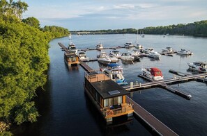 Un port de plaisance avec des bateaux amarrés, une structure flottante dotée d’une terrasse et une rive bordée d’arbres.