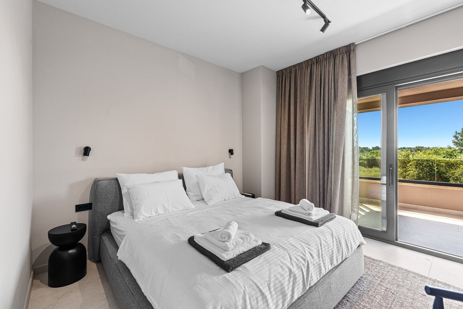 A modern hotel room with a large bed, a small side table, a glass door leading to a balcony, and a view of greenery.