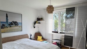 Une chambre avec un grand lit, une chaise, une fenêtre avec des stores, un radiateur et un tableau accroché au mur.