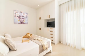 Un dormitorio con cama, televisor, un cuadro y ventana con cortinas.