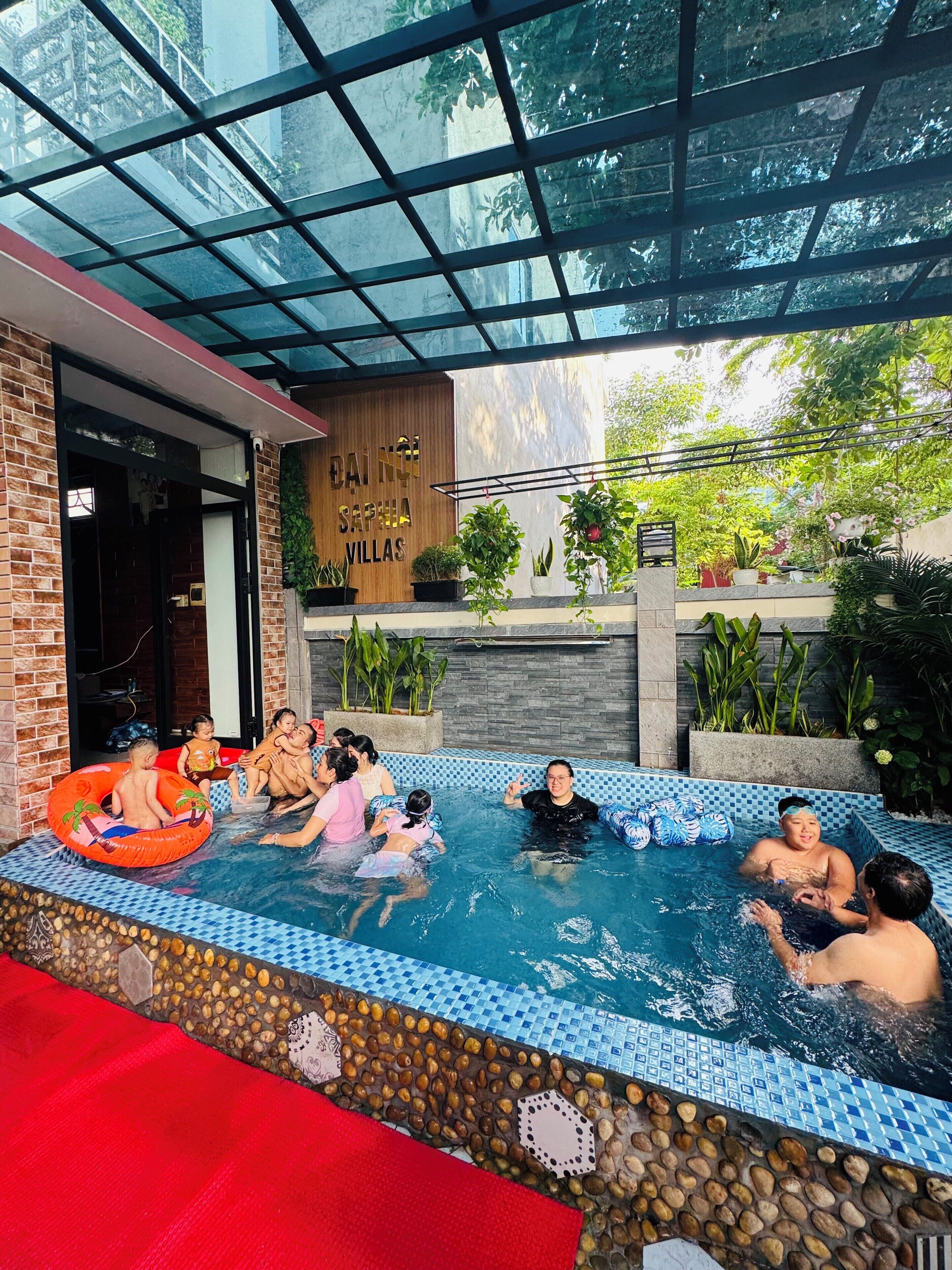 Zona de piscina exterior con techo de cristal, rodeada de plantas y un letrero de madera que dice 'DAI NOI SAPPHIA VILLAS'.
