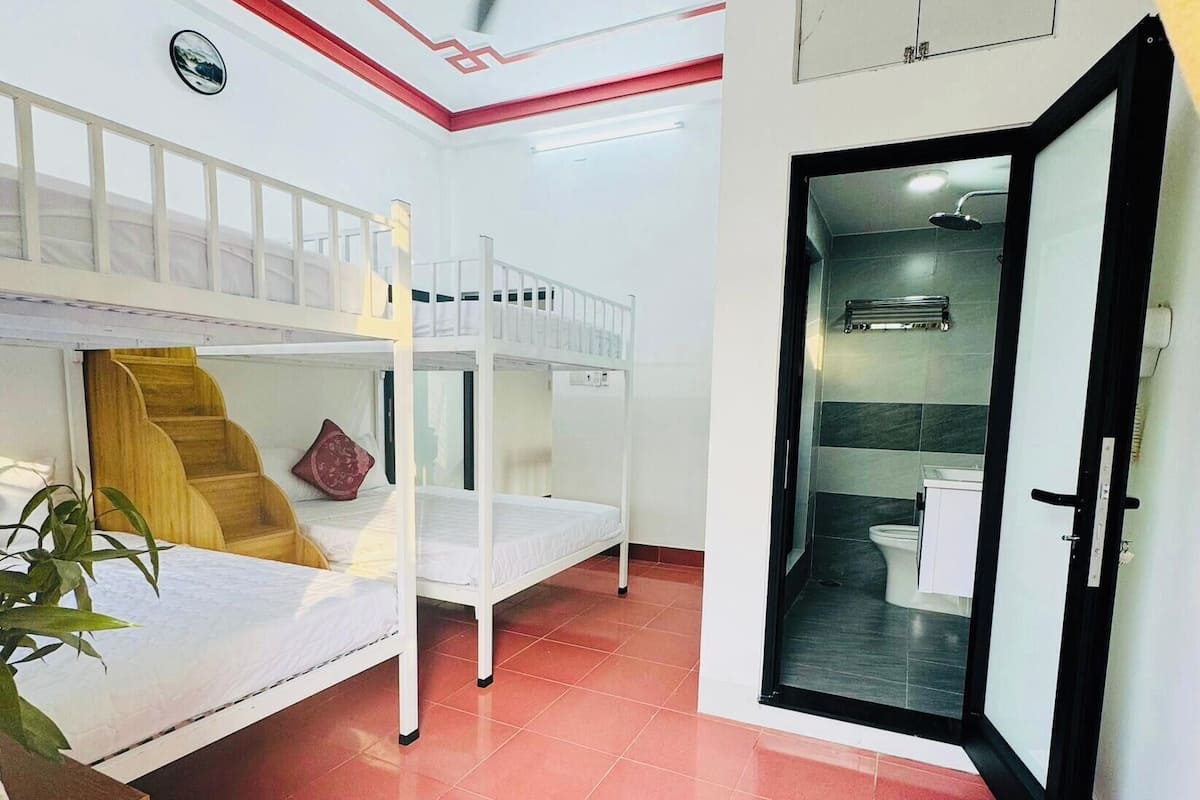 A dormitory room with bunk beds, a ceiling fan, a bathroom with a toilet and sink, and a door leading to another room.