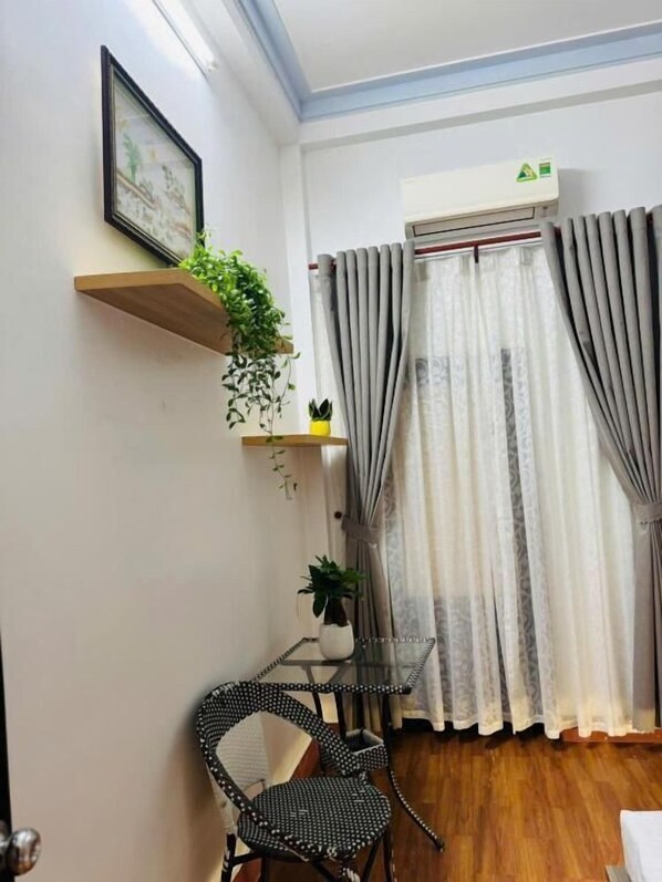 A room with a wooden floor, a black chair, a small table with a plant, a framed picture on the wall, and a window with grey curtains.