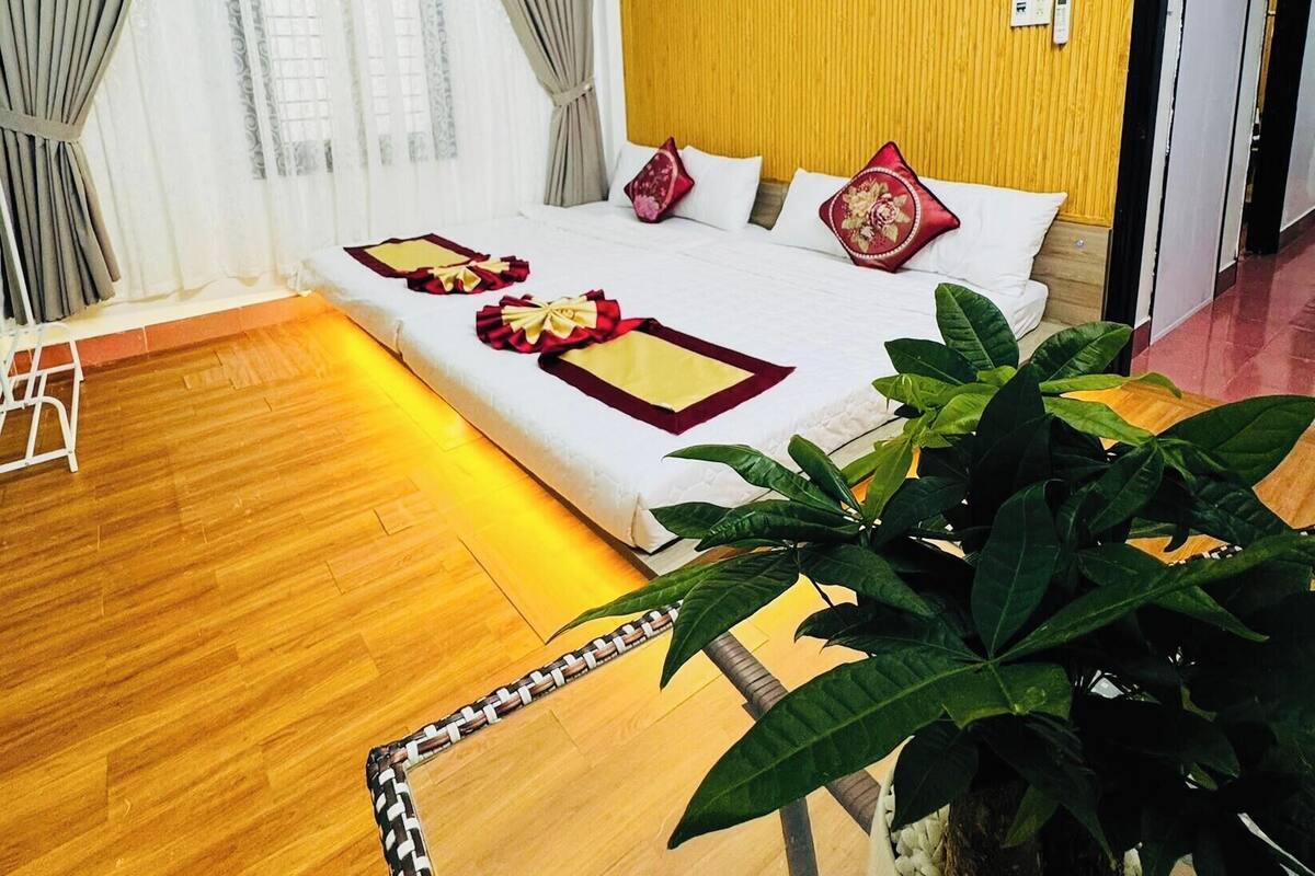 A hotel room with a wooden floor, a bed with red and white bedding, a large potted plant, and a yellow wall.
