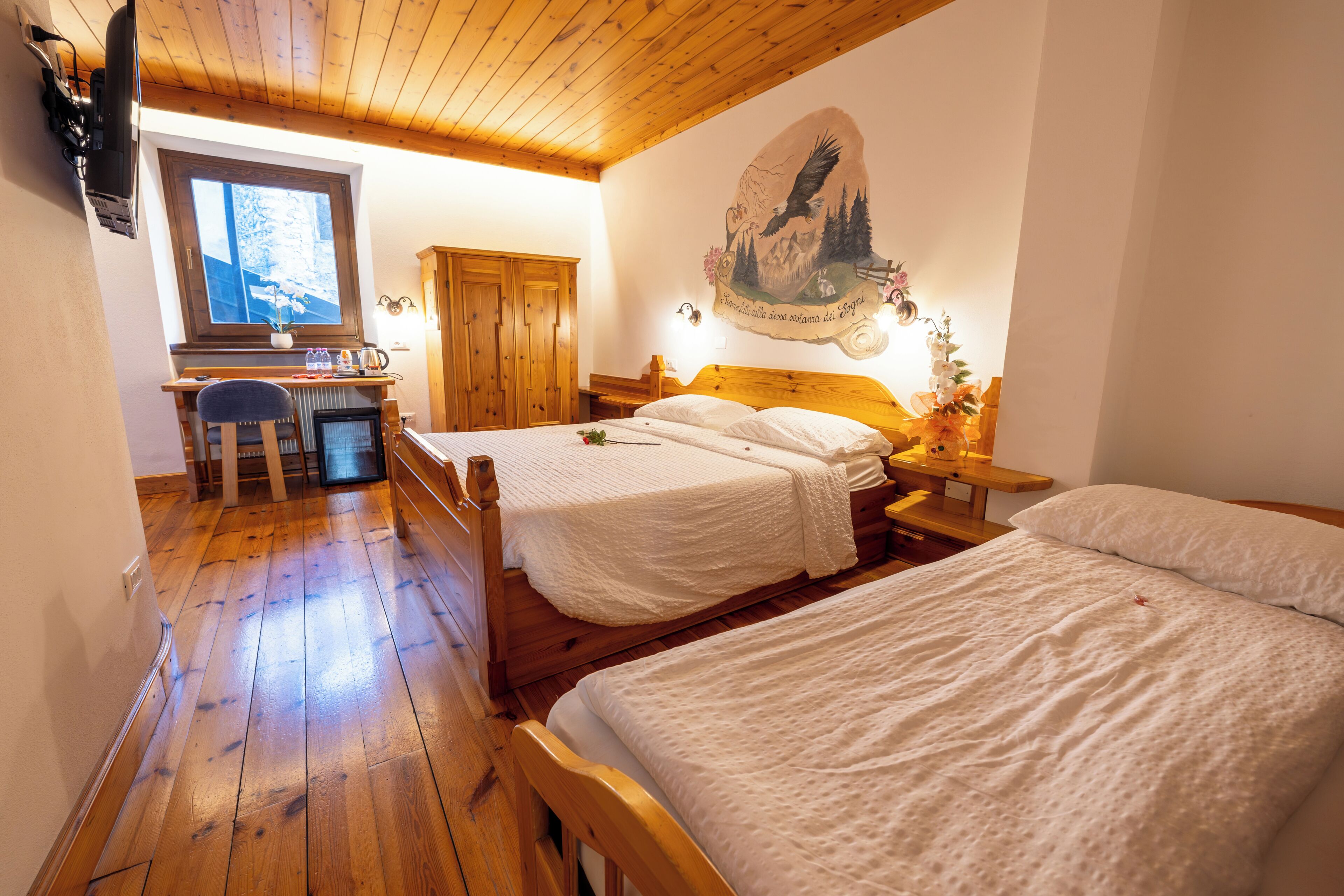 A bedroom with two beds, wooden flooring, a desk, a chair, and a wall-mounted TV.