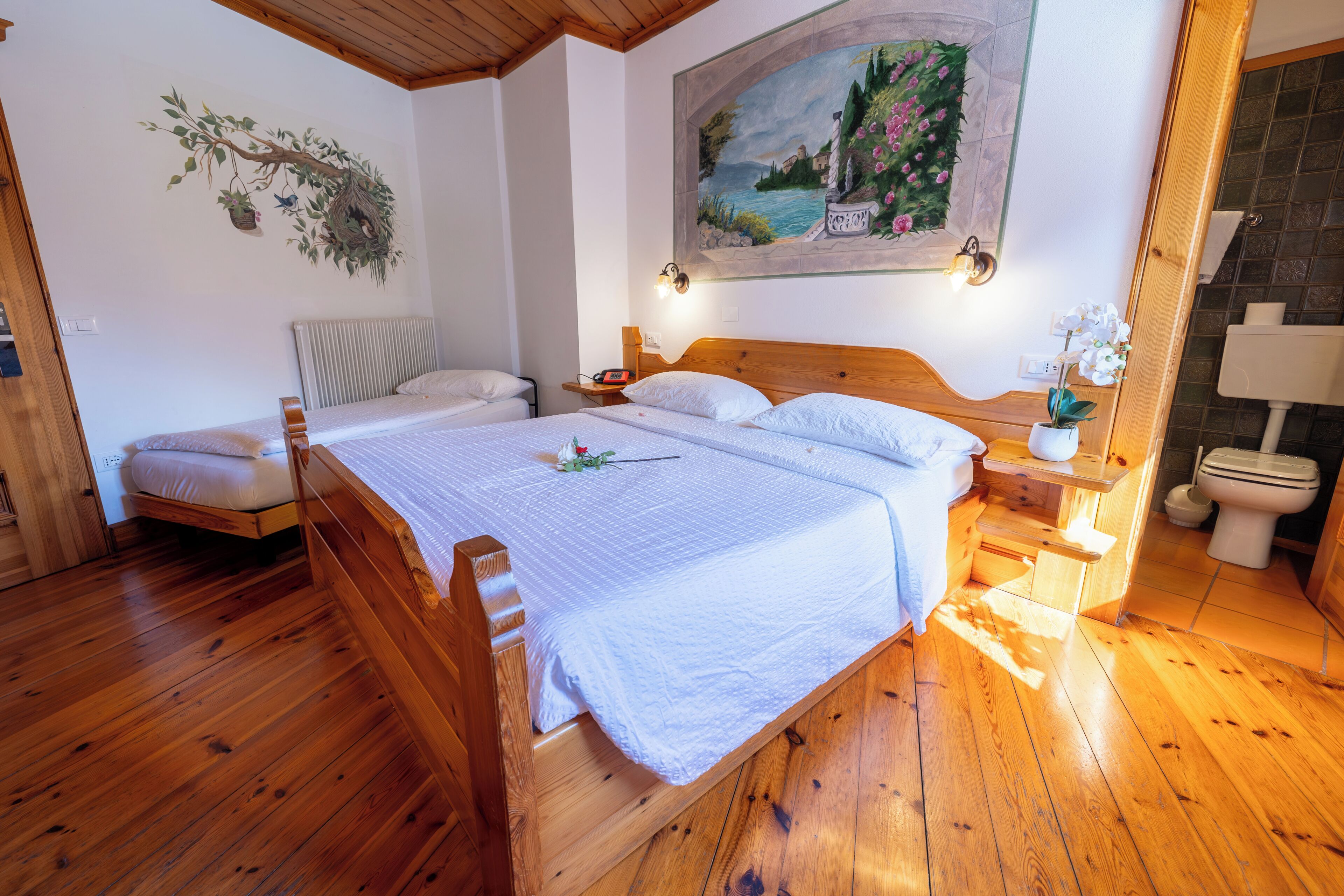 Un dormitorio con piso de madera, una cama doble, una mesita de noche, un cuadro en la pared y un baño con inodoro y lavamanos.