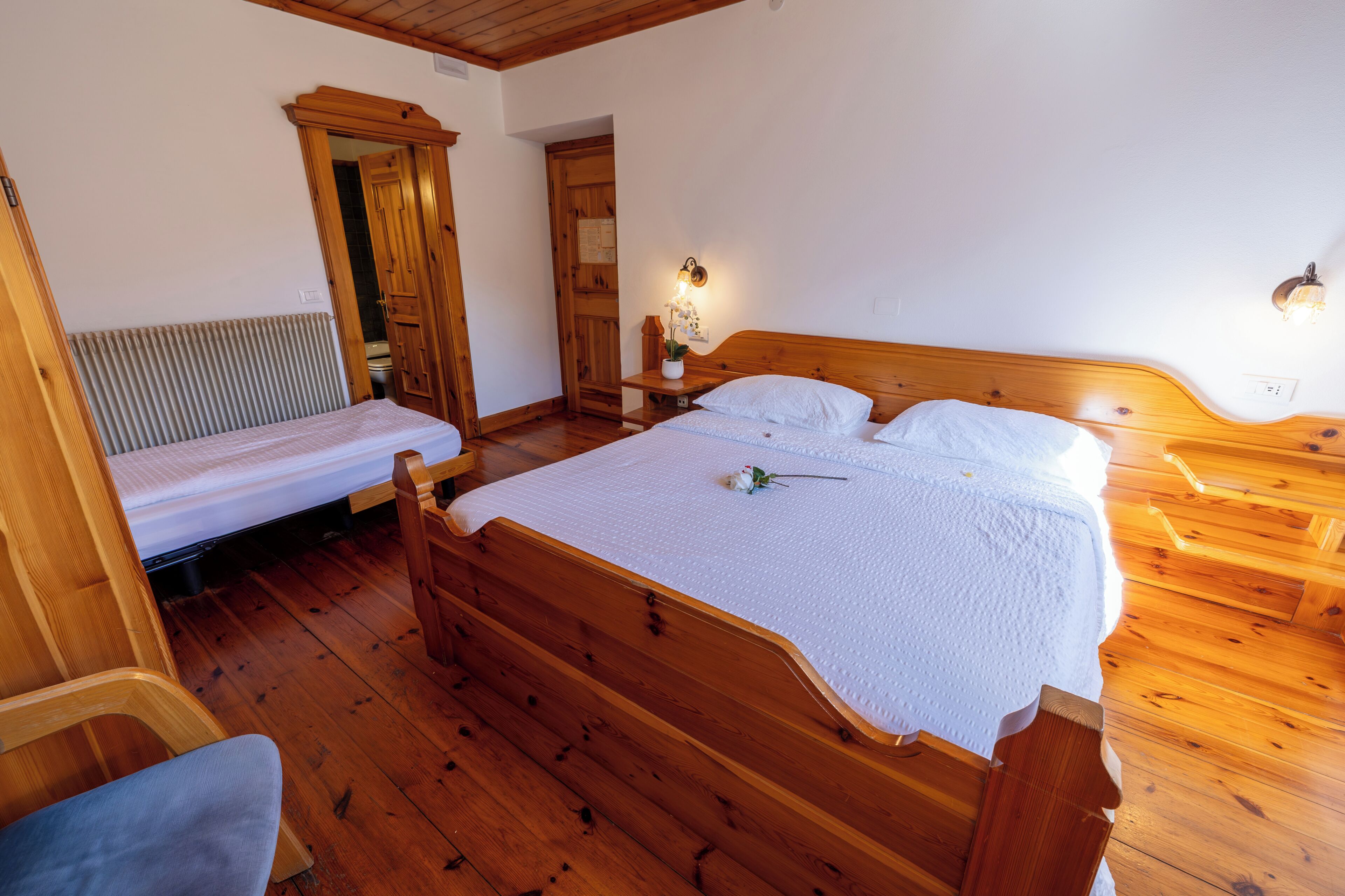 Una cama de madera con colcha blanca, una mesita de noche de madera con lámpara y una silla de madera.