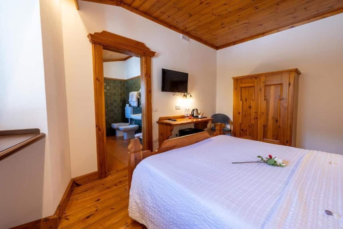 A bedroom with a wooden ceiling, a bed with a white bedspread, a wooden desk with a chair, a television mounted on the wall, and a wooden wardrobe.