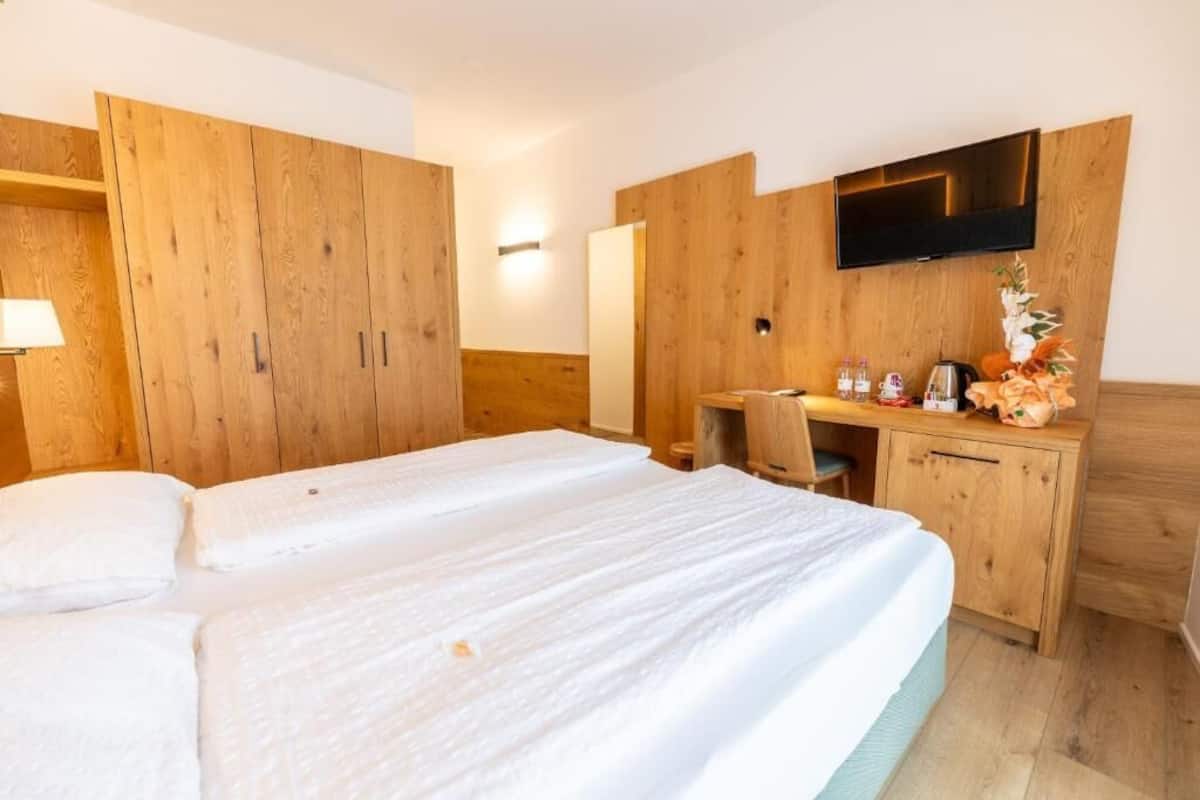 A hotel room with a bed, wooden wardrobe, desk, chair, television, and a small table with toiletries.