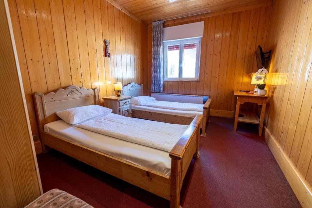 Ein Zimmer aus Holz mit zwei Betten, einem Fenster mit Vorhängen, einem Nachttisch mit Lampe und einem kleinen Tisch mit Pflanze.