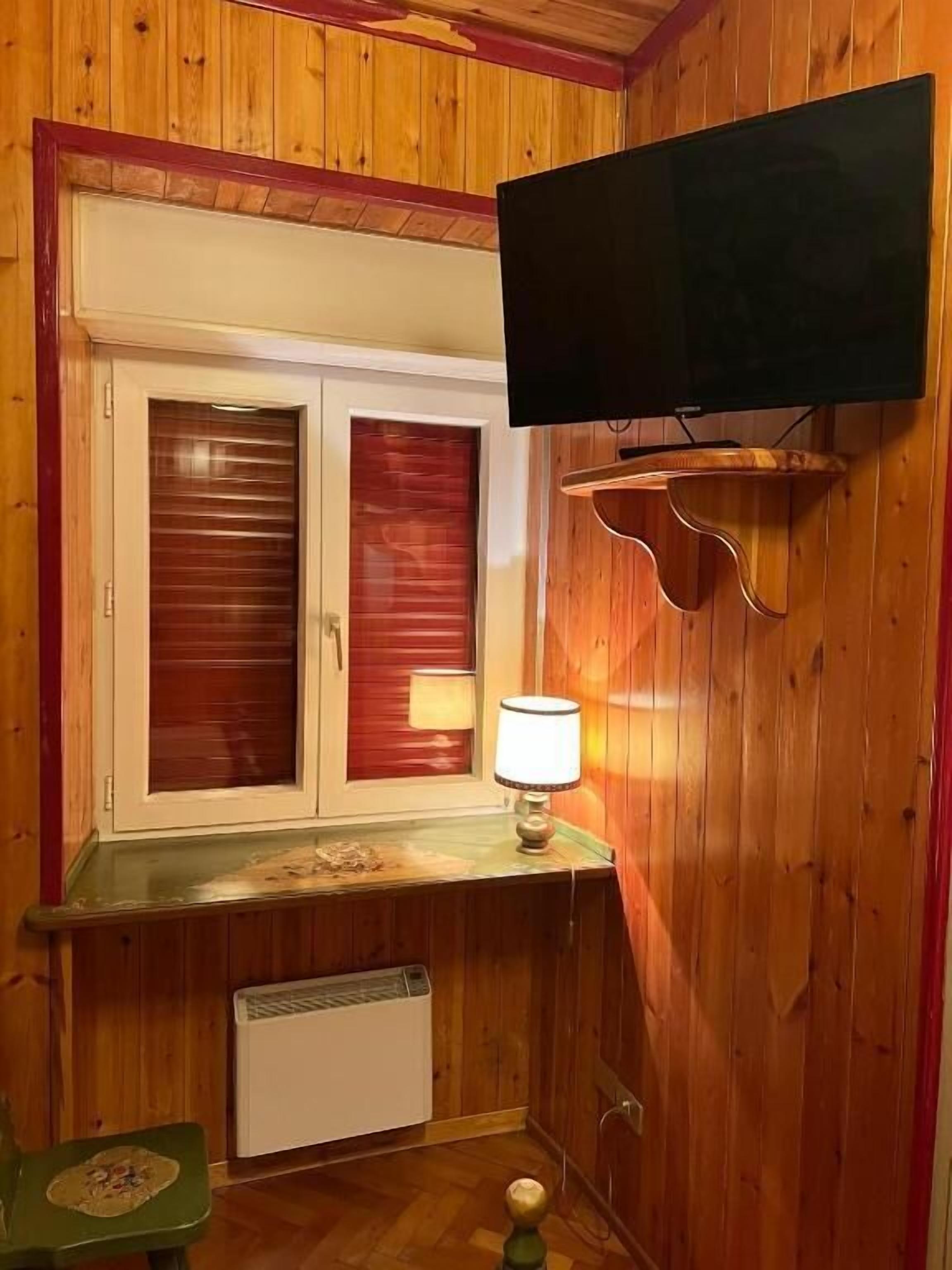 A room with wooden paneling, a television mounted on the wall, a window with blinds, a table with a lamp, and a radiator.