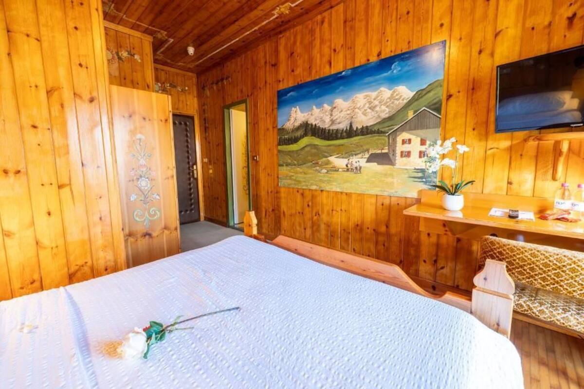 A wooden room with a bed, a painting of mountains, a TV, and a small table with a vase.