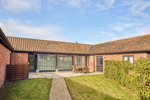 A brick house with a tiled roof, a patio with a table and chairs, and a well-maintained lawn. - The Saddlery (Norwich)