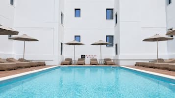 Une piscine entourée de chaises longues et de parasols, elle-même bordée d’un bâtiment blanc doté de nombreuses fenêtres.