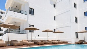 Een zwembadgedeelte met ligbedden en parasols, een helderblauw zwembad en een wit gebouw met balkons.