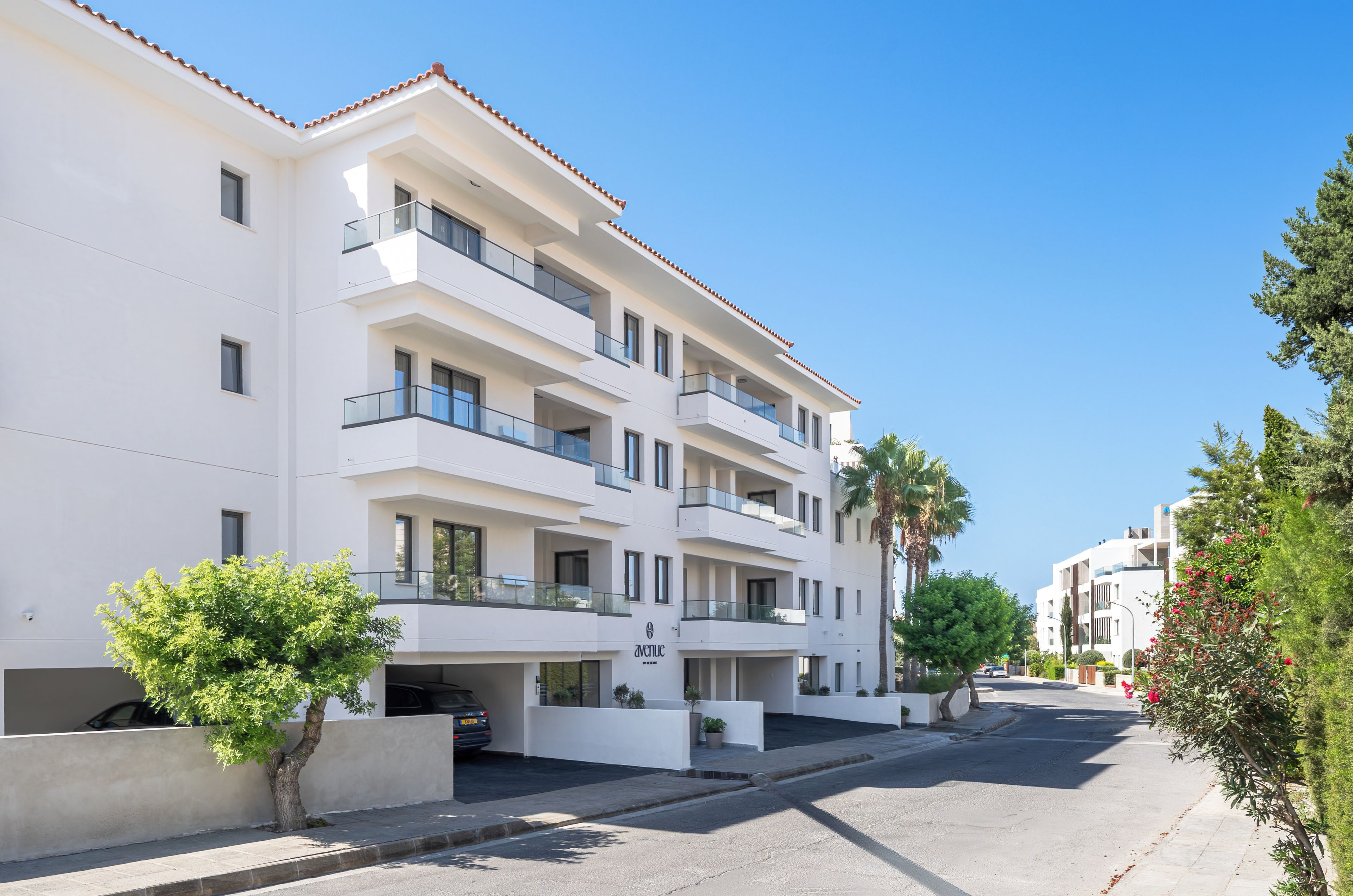 A multi-story residential building with balconies, a parking area, and a clear sky.