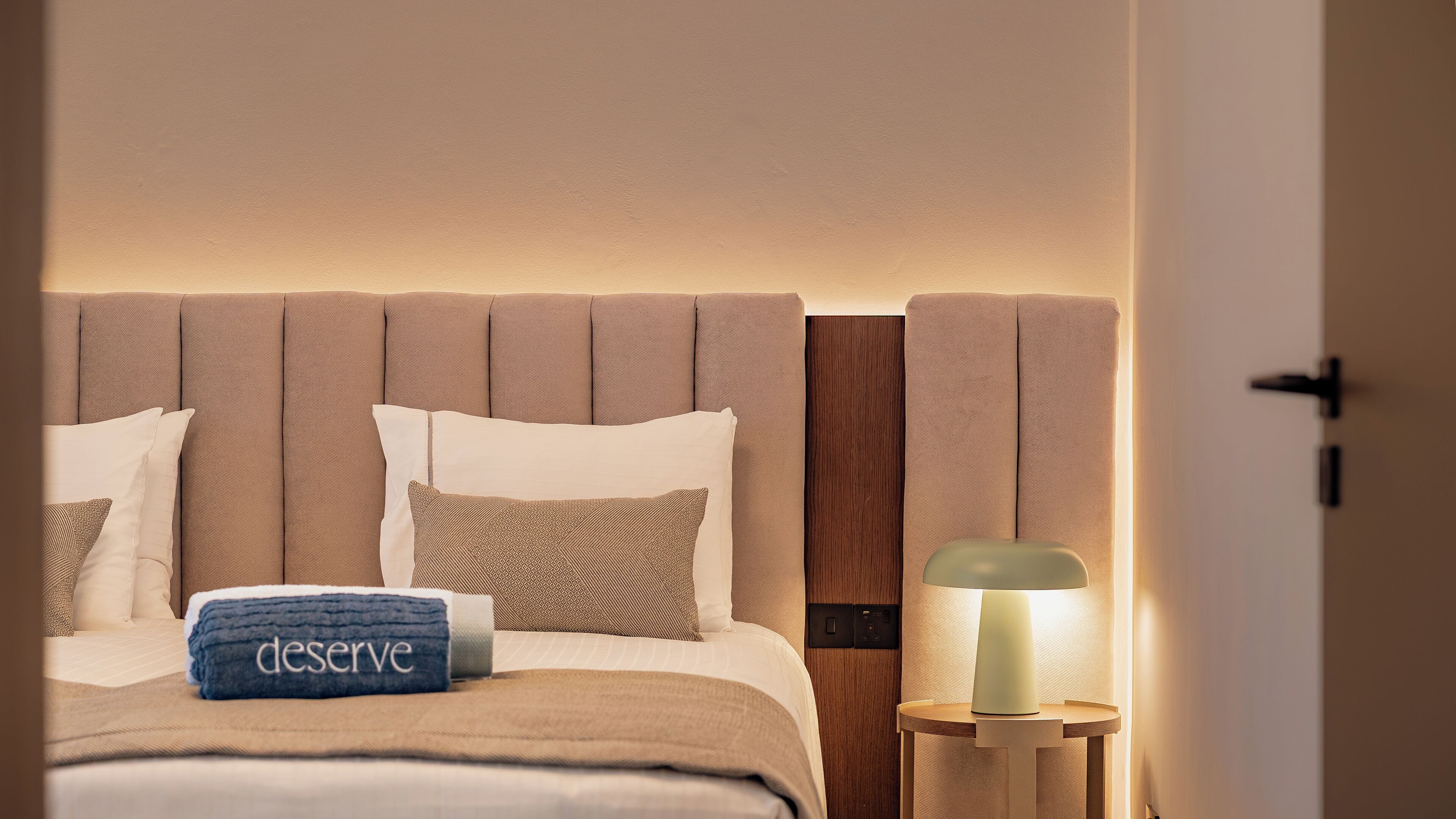 Ein ordentlich bezogenes Bett mit einem blauen Handtuch und einem Kopfkissen mit dem Schriftzug 'deserve', einer Nachttischlampe und einem hölzernen Kopfteil.