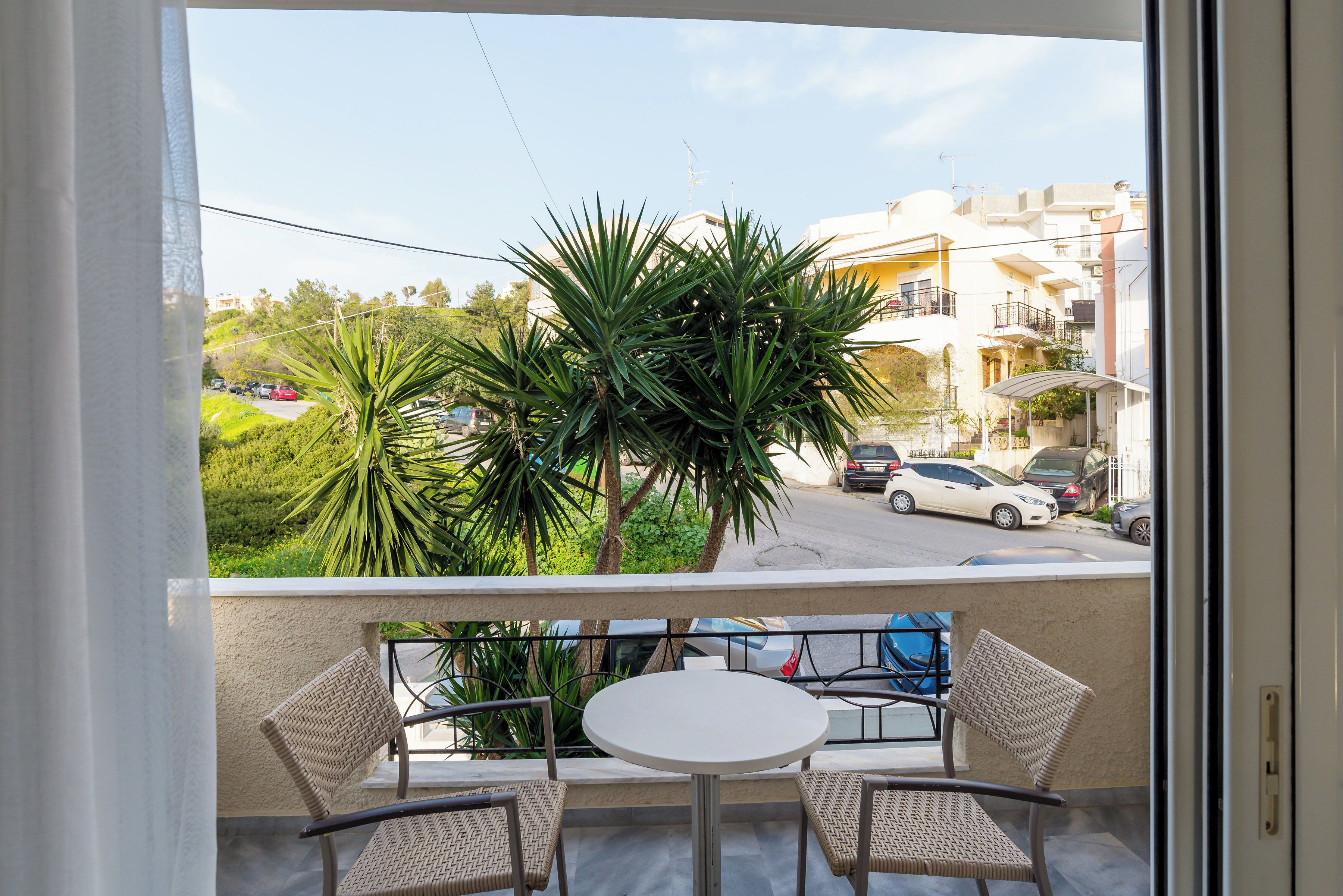 A balcony with wicker chairs and a small table, overlooking a street with parked cars and buildings.