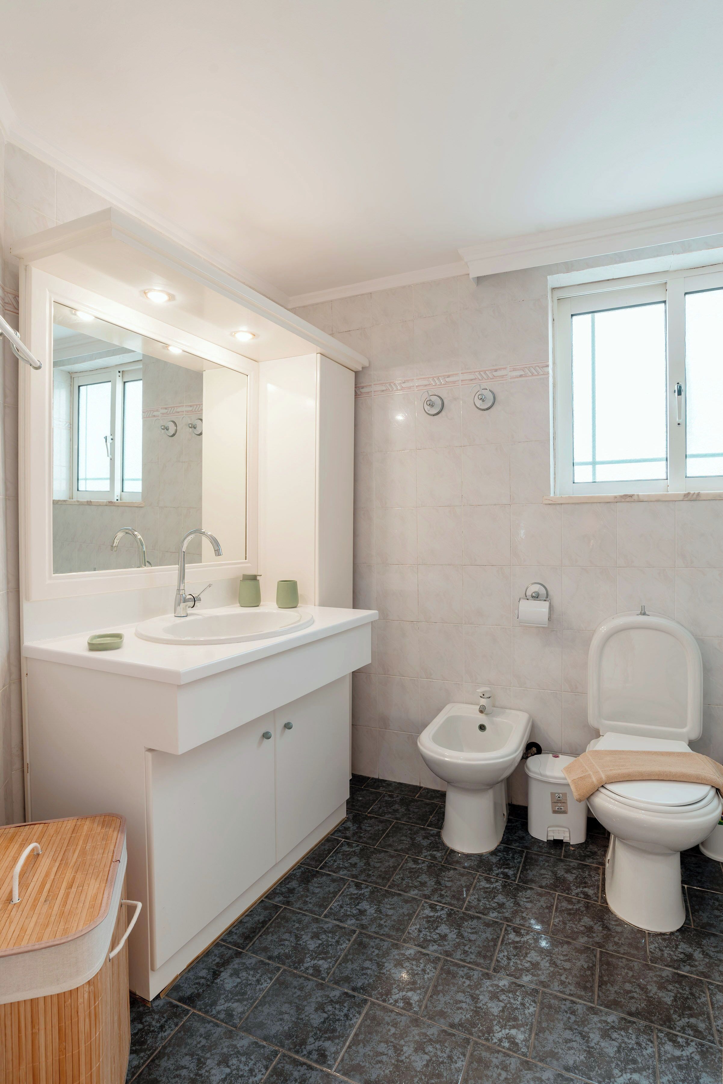A modern bathroom with a sink, bidet, and toilet, a mirror, and a window.