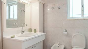 A modern bathroom with a sink, bidet, and toilet, a mirror, and a window.