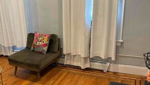 A room with a wooden floor, a grey sofa with a colorful pillow, white curtains, and a black table.