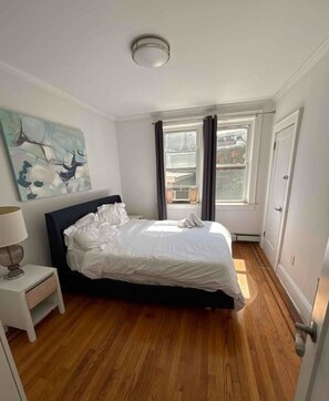 A bedroom with a bed, a nightstand, a painting, a window with curtains, and a door. - Beautiful 2BR/1BA Apt close to NYC! (Jersey City)