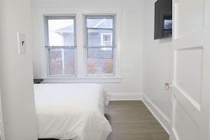 Habitación estrecha con cama, dos ventanas, una puerta y televisor montado en la pared.